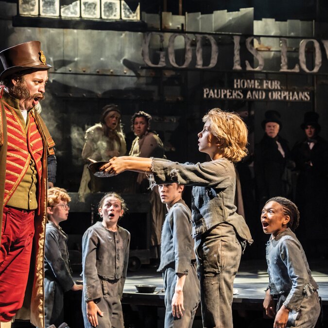 A stage scene showing an adult figure addressing a group of children in worn clothing in front of a large sign on a dimly lit set | © Danny Kaan