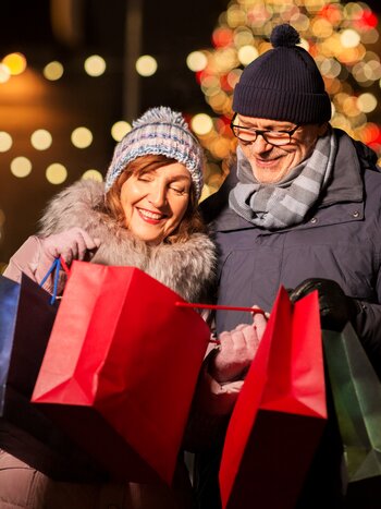 Glückliches Paar mit Einkaufstaschen am Weihnachtsmarkt nach erfolgreichem Weihnachtsshopping-Tag in der festlich geschmückten Stadt
 | © shutterstock / 1494691982