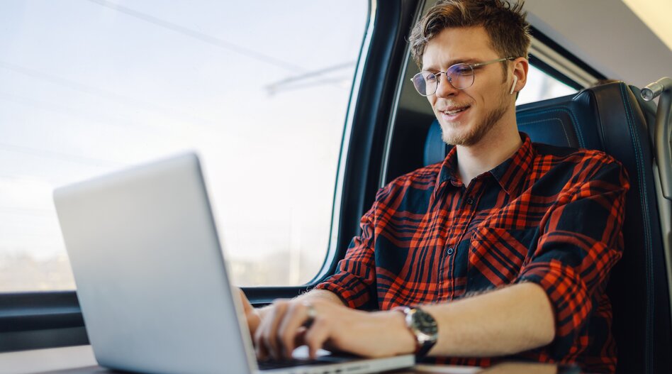 Porträt eines jungen Passagiers, der in der Nähe des Zugfensters sitzt und auf einem Laptop tippt, während er ihn lächelt und mit dem Zug fährt. Ein glücklicher Hipsterreisender arbeitet auf seiner Bahnreise an einem Laptop.