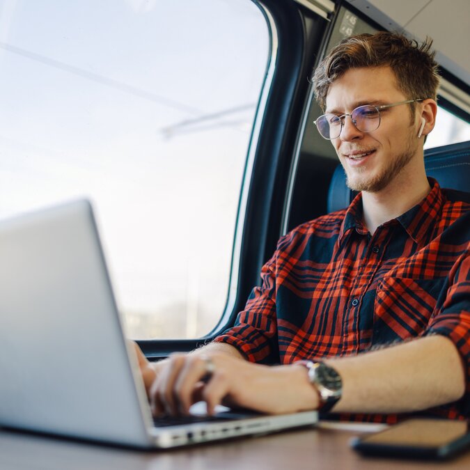 Porträt eines jungen Passagiers, der in der Nähe des Zugfensters sitzt und auf einem Laptop tippt, während er ihn lächelt und mit dem Zug fährt. Ein glücklicher Hipsterreisender arbeitet auf seiner Bahnreise an einem Laptop.