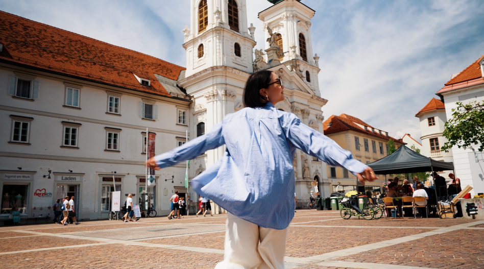 Person mit ausgestreckten Armen bewegt sich über einen sonnigen Platz in Graz, umgeben von historischen Gebäuden, einer barocken Kirche und Marktständen im Hintergrund. | © Österreich Werbung, Stefan Strasser