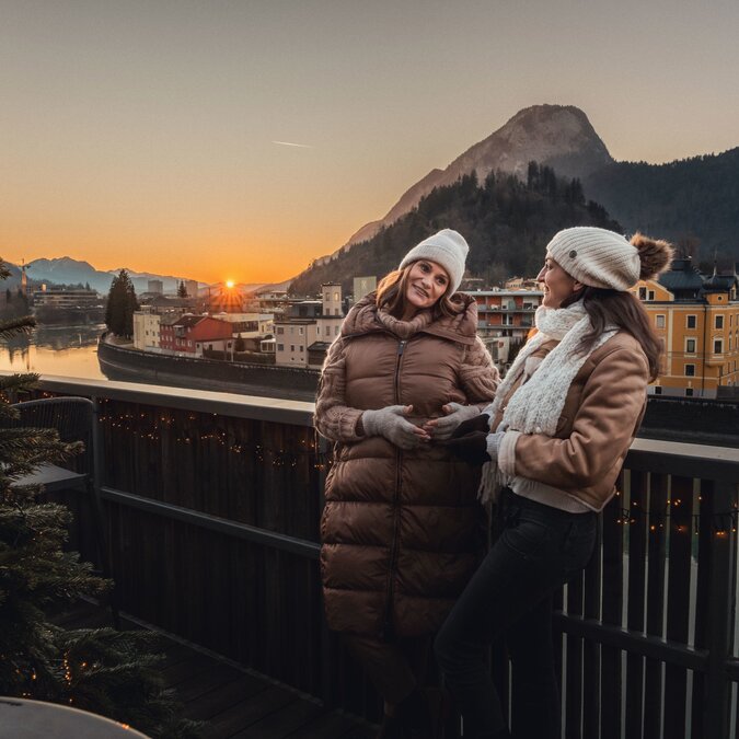 Two people dressed in winter clothing stand on a terrace overlooking a riverside town and the surrounding mountains at sunset. In the foreground, a decorated Christmas tree is illuminated, while the warm evening light highlights the winter landscape. | © TVB Kufsteinerland, Thorben Jureczko