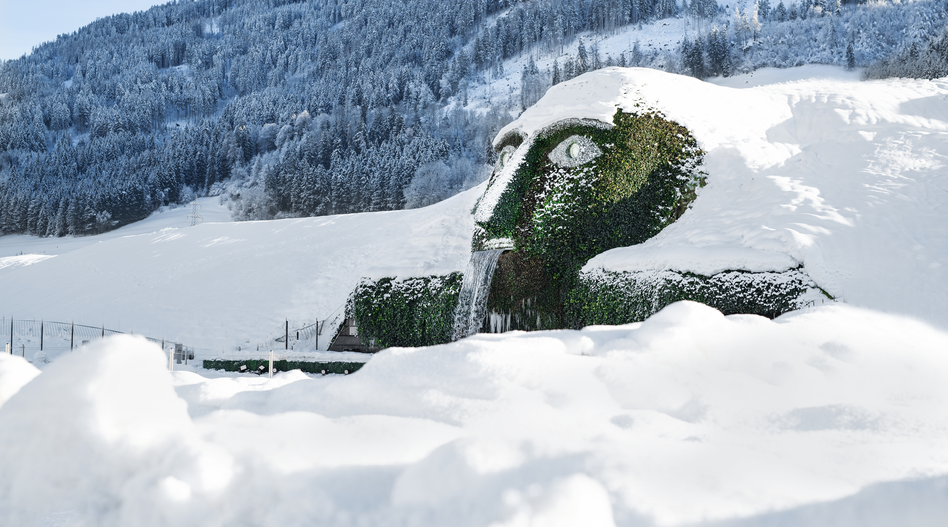 Winterlandschaft mit dem berühmten Swarovski-Riesen in Wattens, dessen mit Pflanzen bedecktes Gesicht teilweise von Schnee bedeckt ist. Wasser fliesst aus dem Mund der Skulptur, während verschneite Hügel und Wälder die Szene umgeben. | © Swarovski Kristallwelten