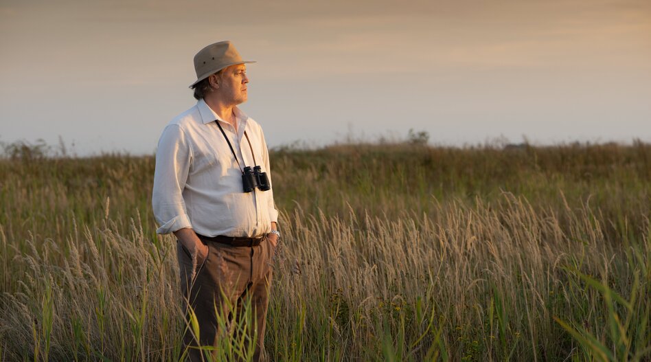 Une personne munie de jumelles se tient dans un vaste paysage de prairies du Burgenland, en Autriche. La lumière chaude du soleil et l’environnement naturel dégagé créent une atmosphère paisible. | © So-Weit-So-Nah-2025_054©Burgenland Tourismus_Andreas Hafenscher