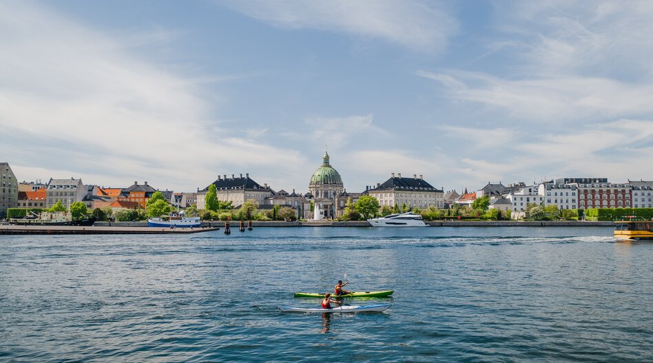 Zwei Kajaks auf einem breiten Kanal vor der Uferpromenade von Kopenhagen mit dem Schloss Amalienborg und seiner Kuppel im Hintergrund unter blauem Himmel. | © Mitch Wiesinger