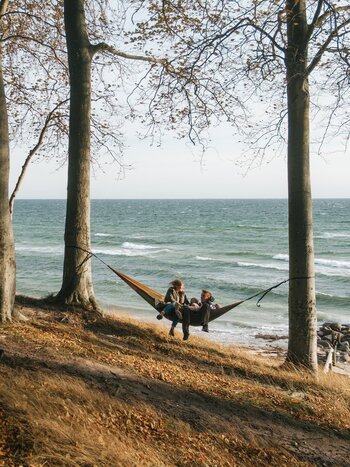 Two people relaxing in a hammock between trees in the coastal forest of Teglværksskoven on the Danish island of Funen. | © Mitch Wiesinger