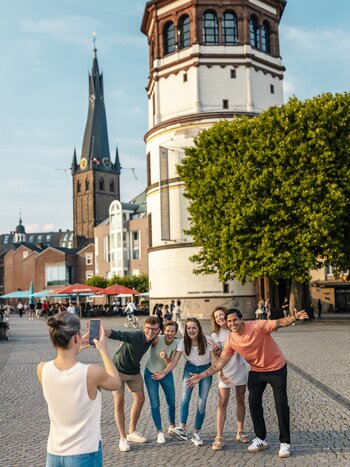 A group takes a photo of themselves in front of the Rheinturm and a church in Düsseldorf on a busy square | © Visit Düsseldorf