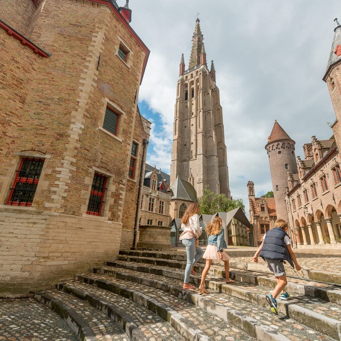 Plusieurs personnes montent un large escalier en pierre dans la vieille ville de Bruges. Elles sont entourées de bâtiments historiques en briques, dont un haut clocher et d'autres parties de bâtiments médiévaux. La place est caractérisée par ses pavés, ses arcades et son architecture typique. | © Jan Darthet 