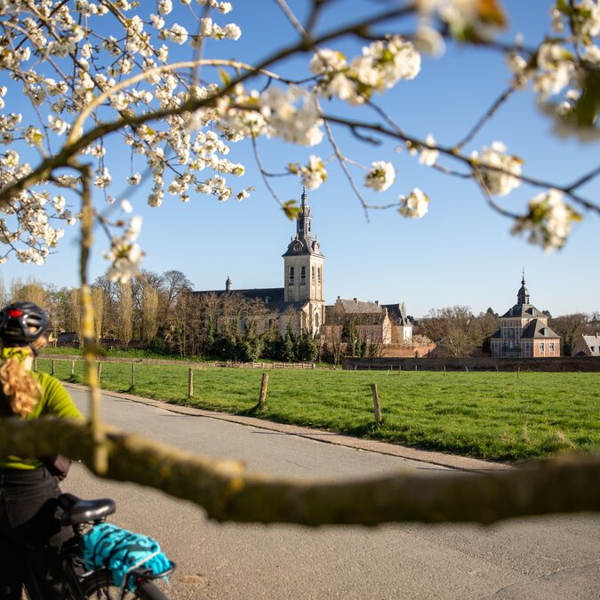 Une personne à vélo sur une route de campagne, devant des arbres fruitiers en fleurs, avec vue sur une église historique et les champs environnants, dans une région rurale. | © Stad Leuven