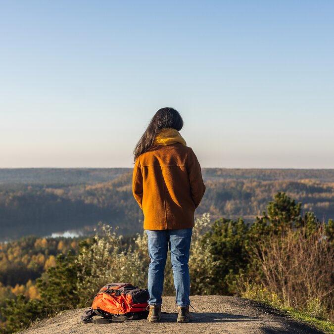 Eine Person steht auf einem Felsvorsprung mit Blick über eine weite Landschaft aus Wäldern und Seen; ein Rucksack liegt am Boden neben der Person. | © MEDIALIFE.BE