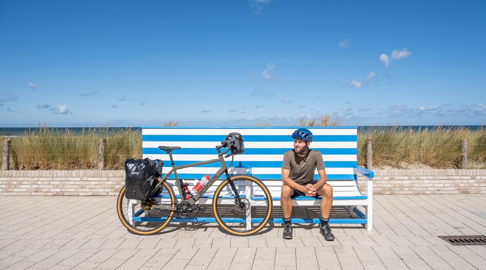 A person is sitting on a blue and white striped bench on a beach promenade next to a fully loaded bicycle. In the background, you can see dune grasses, the sea, and a clear blue sky. | © Bas van Oort