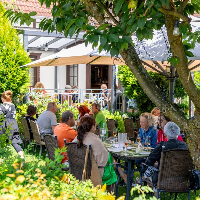 People sit around a table in the green and talk while waiting for their food, in the background a white and brown house. | © #visitrheinmain | David Vasicek