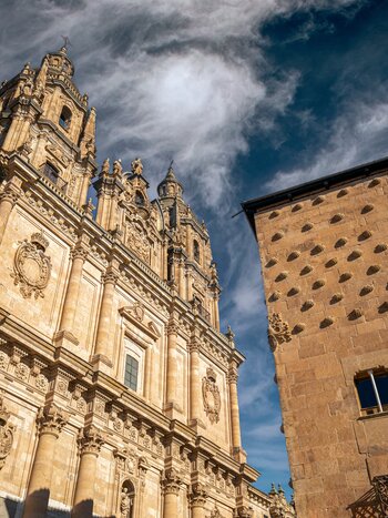 Casa de las Conchas in Salamanca, Spain, with its distinctive shell-covered façade beside a Baroque church and the historic old town under a dramatic sky. | © Turespaña