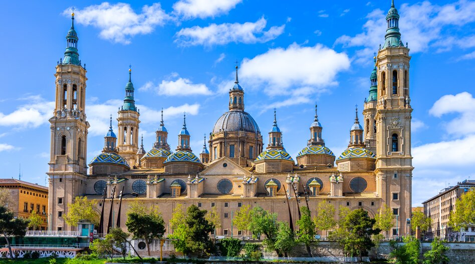 Basílica del Pilar in Saragossa in Spanien mit markanten Türmen, kunstvollen Kuppeln und prachtvoller Barockarchitektur am Ufer des Ebro unter blauem Himmel. | © Turespaña