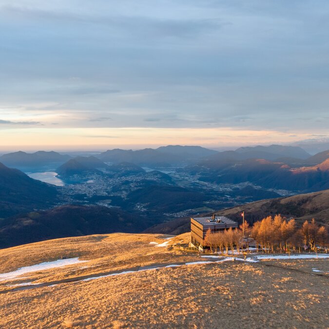 Large panorama depuis la Capanna Monte Bar sur la région de Lugano avec collines, montagnes et le lac de Lugano baignés de lumière dorée. | ©  Lugano Region-DigitalFlow.ch