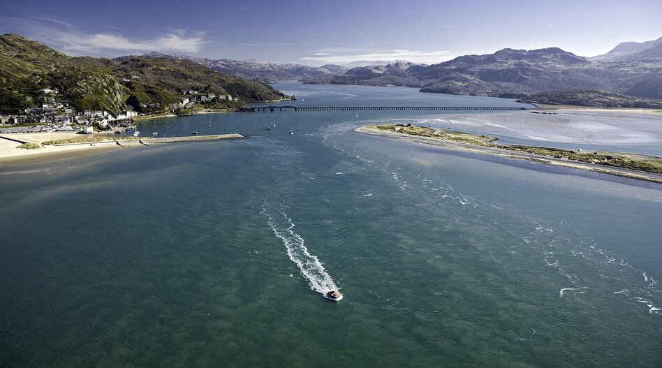 Luftaufnahme der Mawddach-Mündung bei Barmouth in Wales mit der historischen Barmouth Bridge, Sandbänken, Boot und den Bergen des Snowdonia-Nationalparks im Hintergrund. | © Crown Copyright (2026) Welsh Government