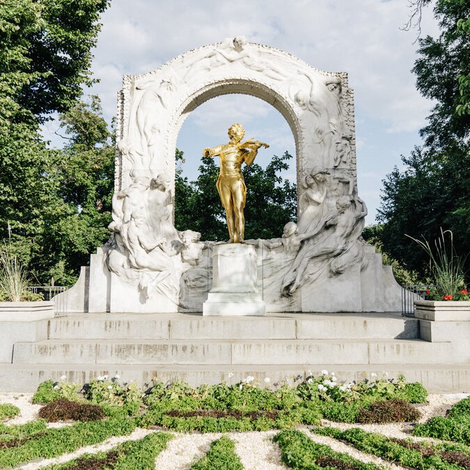 Die goldene Statue von Johann Strauss II steht im Wiener Stadtpark unter einem kunstvoll verzierten weißen Marmor-Bogen. | © Wien Tourismus | Gregor Hofbauer