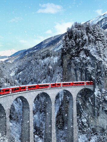 The Bernina Express crossing the snow-covered Landwasser Viaduct in Switzerland during winter, surrounded by snowy mountains and forests. | © shutterstock / 2395790421