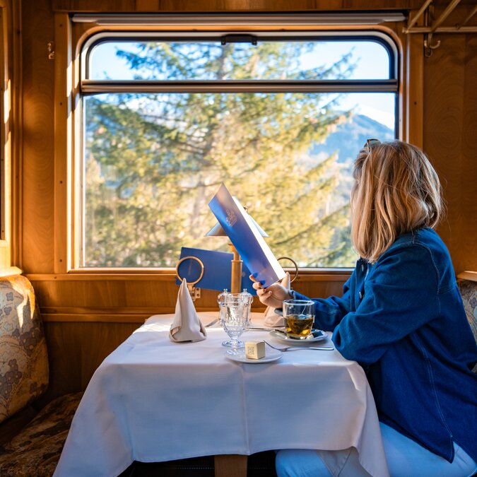 Une personne est assise dans le Gourmino, la voiture-restaurant du Glacier Pullmann Express, et regarde par la fenêtre. Elle tient un menu dans la main et un thé est posé devant elle. | © Rhätische Bahn 