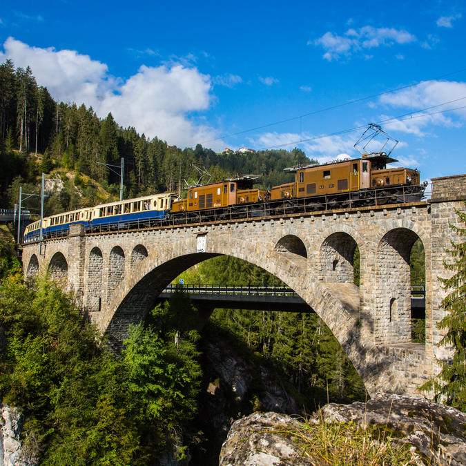 Der Glacier Pullmann Express mit einer braunen Lokomotive und weiss-blauen Wagen fährt über eine Brücke mitten im Wald, dahinter die Berge und ein blauer Himmel. | © Rhätische Bahn 
