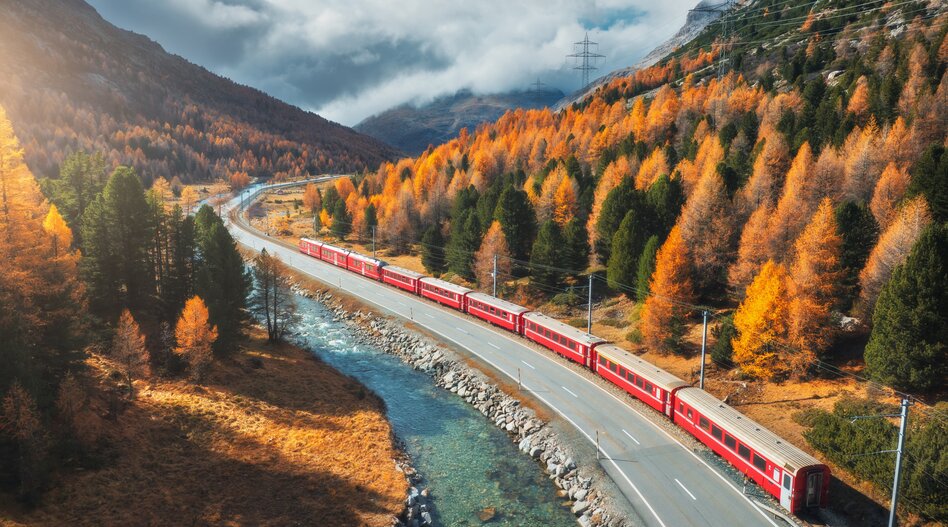 Roter Zug fährt entlang eines Flusses durch herbstliche Alpenlandschaft. | © shutterstock_2536855561