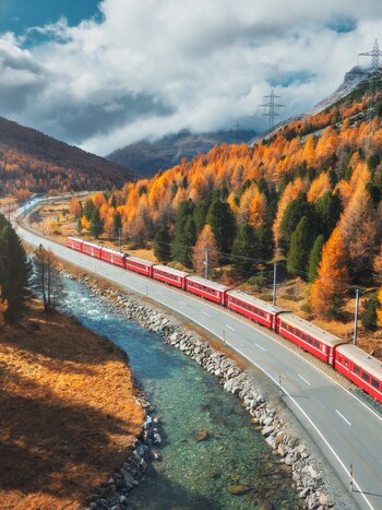 Red train travelling alongside a river through an autumn alpine landscape. | © shutterstock_2536855561
