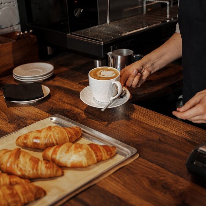 Un barista place un cappuccino avec latte art à côté de croissants fraîchement sortis du four sur un comptoir en bois dans une ambiance de café confortable