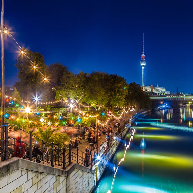 People dancing at summer beach bar beach party near Spree river on historic Museum Island with famous TV tower in the background during blue hour at night, Berlin Mitte district, Germany