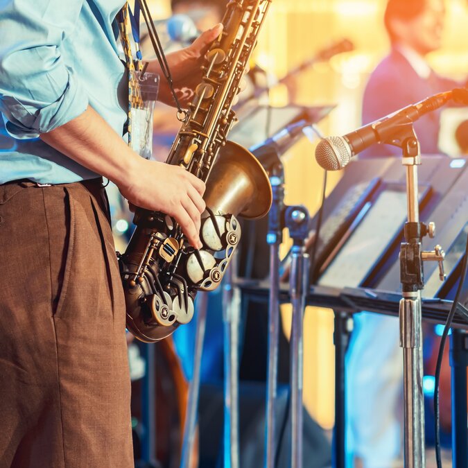 Musician playing saxophone on blurred background. Man with friends blowing saxophone with band for performance. Musical instrument played by a saxophonist musician in festival. | © Shutterstock 2467859293