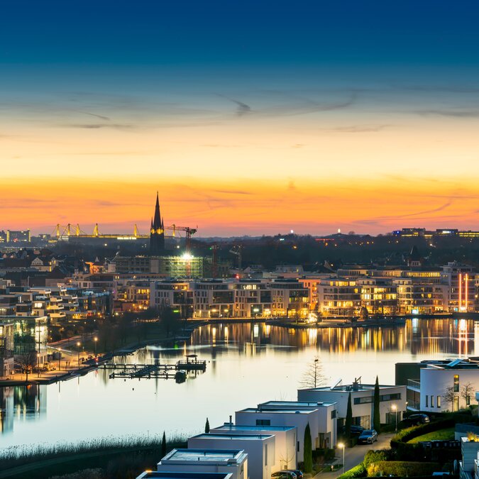 Blick auf den Dortmunder Phoenix-See mit beleuchteter Skyline und Florianturm bei Sonnenuntergang
 | © shutterstock_2166007865