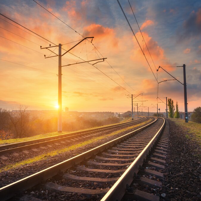 Gare contre beau ciel au coucher du soleil Paysage industriel avec chemin de fer, ciel bleu coloré avec nuages rouges, soleil, arbres et herbe verte | © Shuterstock 513158971