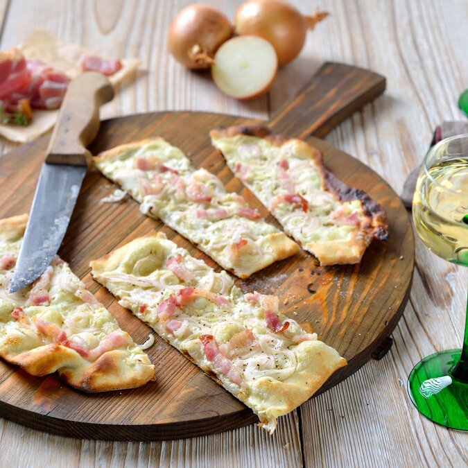 Typischer Flammkuchen aus dem Elsass mit saurer Sahne, Zwiebeln und geräuchertem Speck, heiß aus dem Ofen, serviert mit elsässischem Weißwein | © shutterstock 2060652770