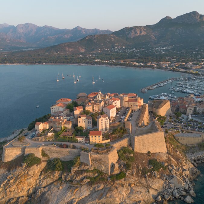 Aerial view of the port town of Calvi in Corsica, featuring an impressive citadel, a marina and the turquoise-blue Mediterranean Sea set against a mountainous coastal landscape. | © shutterstock 2497362275