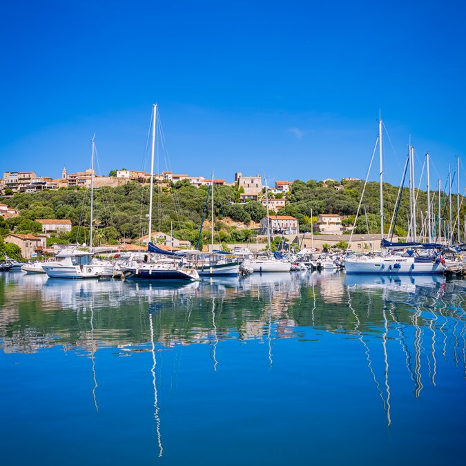 An idyllic marina in Porto-Vecchio, Corsica, with sailing boats, calm waters, a Mediterranean village and a bright blue sky. | © shutterstock 2758010629