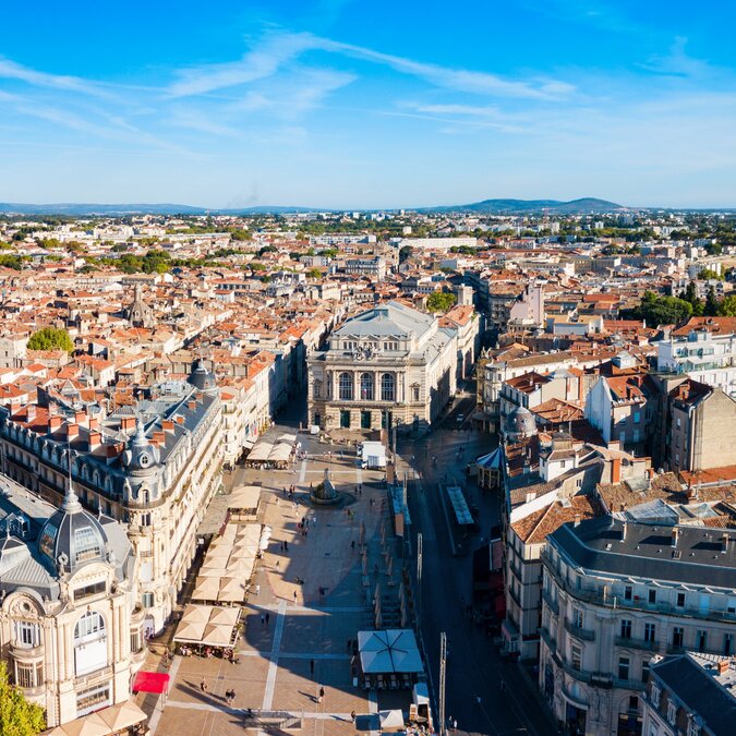 Blick von oben auf den belebten Place de Comédie, gesäumt von den Häusern Montpelliers bei strahlendem Sonnenschein. | © shutterstock / 2423734755