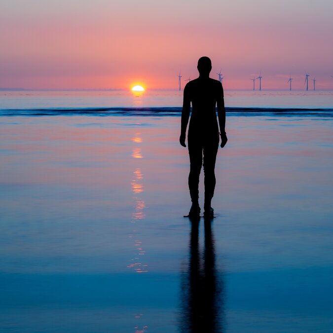 Statue einer Person im Sand mit Blick auf den Sonnenuntergang | © shutterstock / 2351447715