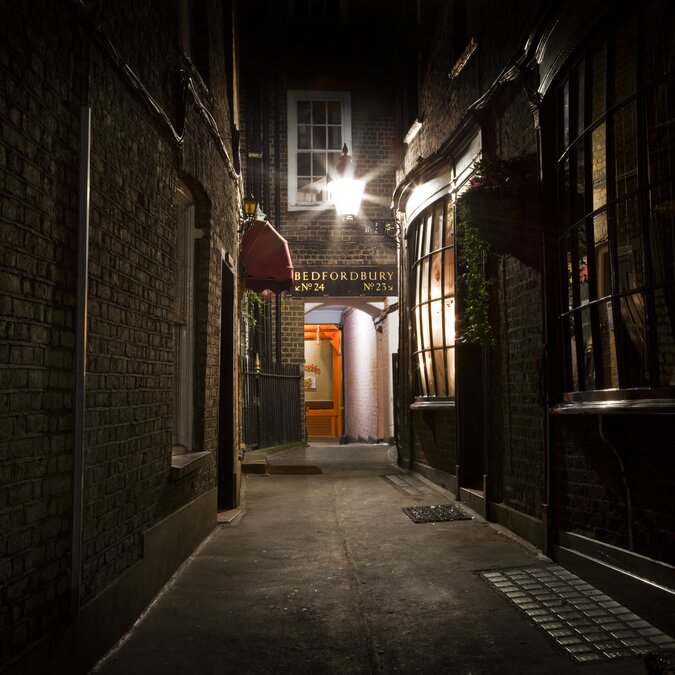 Eine enge, dunkle Gasse mit Backsteinmauern und einem beleuchteten Eingangsschild mit der Aufschrift "Bedfordbury". | © shutterstock // 125129336