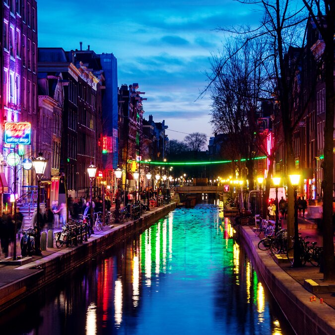 Amsterdam, Pays-Bas. Vue nocturne sur le rouge - phare à Amsterdam, Pays-Bas avec boutiques, clubs de strip-tease, théâtres pour adultes, etc. Coucher de soleil avec reflet dans l'eau | © Shutterstock 1419933056