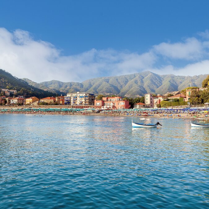 Küste von Levanto mit bunten Häusern, Bergen im Hintergrund und kleinen Booten auf ruhigem Meer.
 | © shutterstock_2192962987