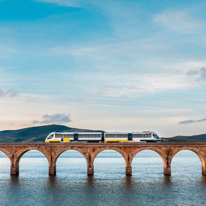 Zugreise durch Spanien mit modernem Zug auf historischer Steinbrücke über Wasser, weiter Landschaft und ruhiger Stimmung unter hellem Himmel. | © shutterstock 1378378817
