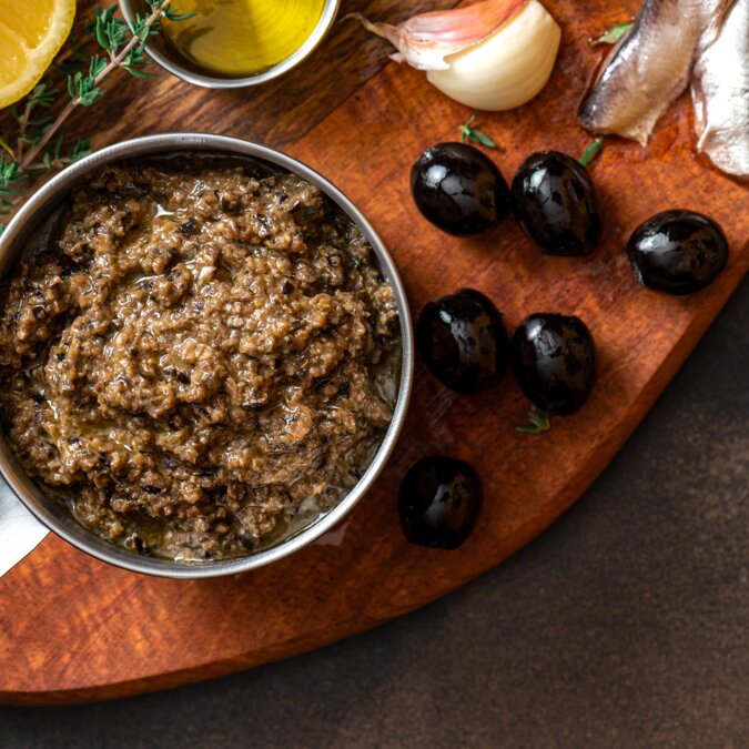 Tapenade in einer Schüssel, Nahaufnahme und Zutaten für Tapenade. Französische Küche. Tapenade wird aus Oliven, Kapern, Sardellen und Kräutern hergestellt. | © Shutterstock 2209498627